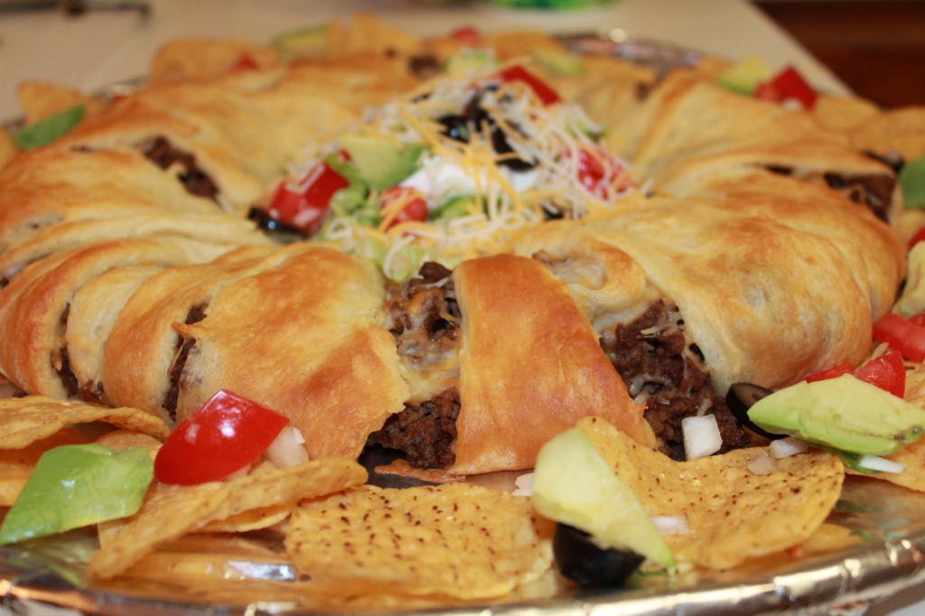 A golden crescent roll taco ring on a foil-lined baking sheet. Tortilla chips and veggies lay around the outside.