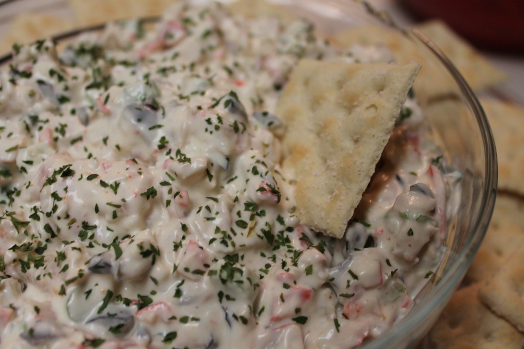 A large bowl of creamy seafood salad is shown with a single saltine cracker dipped into the bowl. The creamy salad is topped with dried parsley flakes.