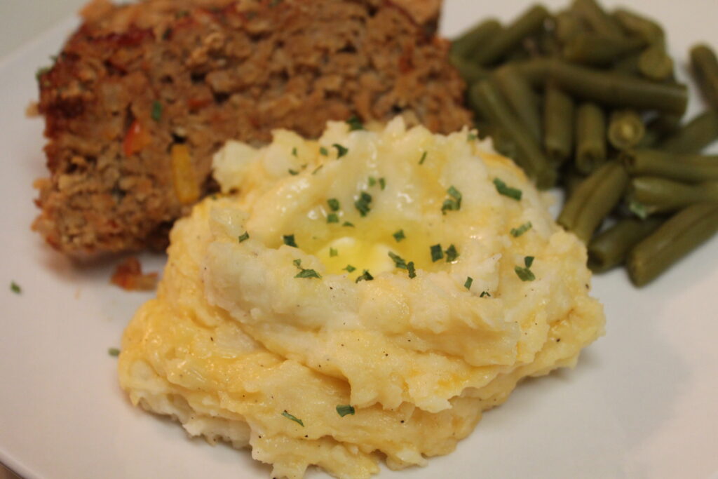 Cheddar garlic mashed potatoes served on a plate alongside a slice of meatloaf and green beans. The mashed potatoes are topped with fresh chives and melted butter.