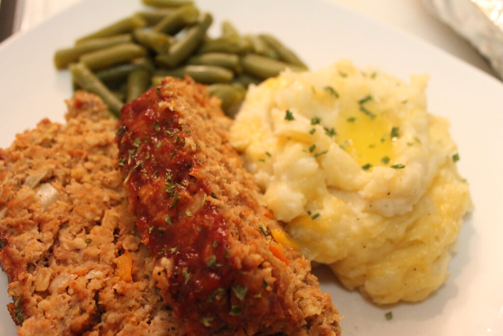 Two slices of meatloaf served with green beans and cheddar garlic mashed potatoes. The creamy mashed potatoes are garnished with melted butter and fresh chives.