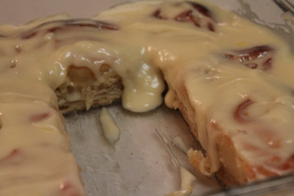 A silver pan full of golden sweet orange rolls with homemade cream cheese frosting running down the side. One roll is missing from the middle. 