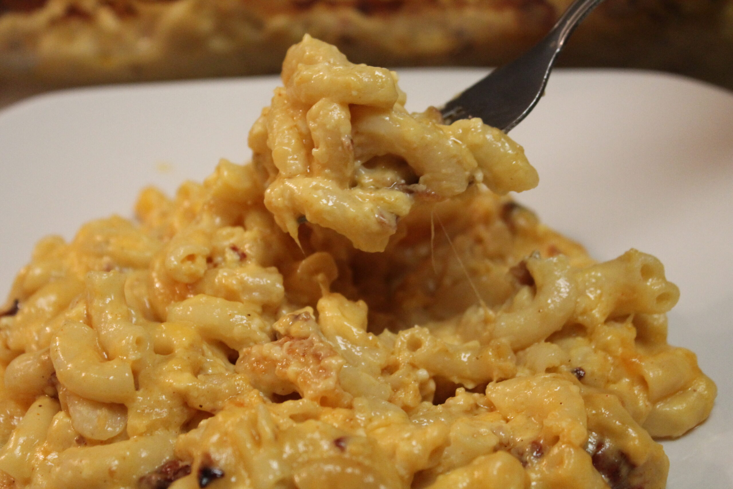 A large helping of creamy bacon mac and cheese is served on a white plate. A silver fork holds a bite of baked mac and cheese.