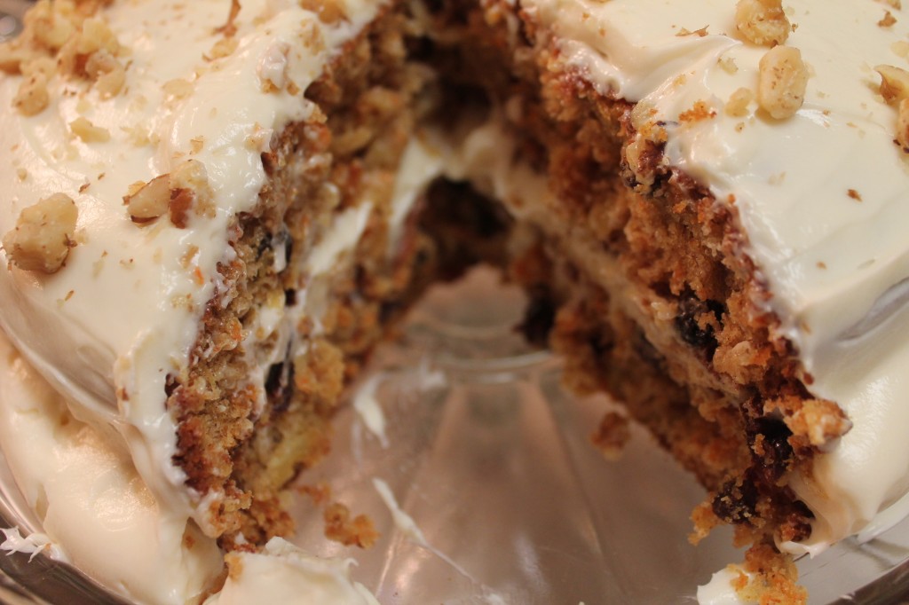 Two layers of ultimate carrot cake slathered in a sweet cream cheese frosting and served on a clear cake dish. Raisins and walnuts are scattered throughout.