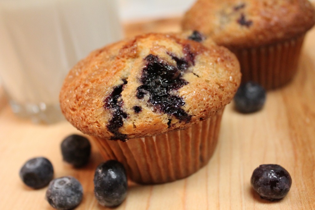 Two warm and fluffy blueberry muffins filled with cheesecake pieces and topped with sugar sitting on a wooden cutting board. Fresh blueberries are scattered nearby.  