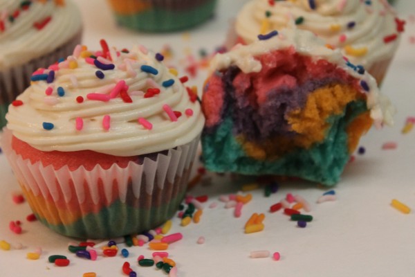 Rainbow cupcakes with vanilla frosting and colorful sprinkles on a white table. One cupcake is halved, showing bright layers of color. 