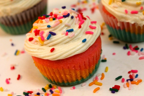 Rainbow cupcakes topped with a swirl of vanilla frosting and sprinkled with rainbow candies. 