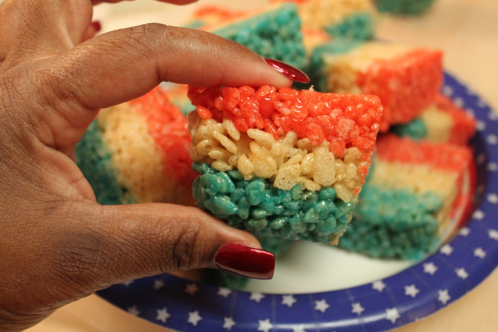 Rosie holds up a square of red, white, and blue layered rice krispies treats, with a plate full of rice krispies squares in the background.