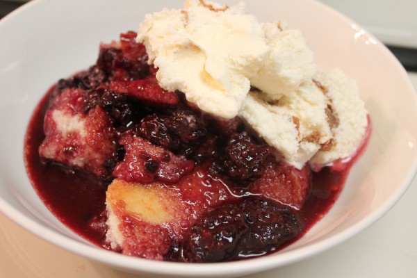 A white bowl filled with homemade blackberry cobbler topped with a scoop of creamy vanilla ice cream.