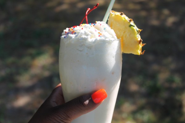 A hand with coral-colored nail polish holds a creamy pineapple and passion fruit milkshake in a tall clear glass. It's garnished with rainbow sprinkles, cherries and pineapple. 