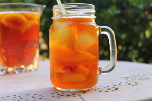 Golden sweet peach iced tea in a clear mason jar glass set on an outdoor patio table. There is a clear pitcher of tea in the background. 