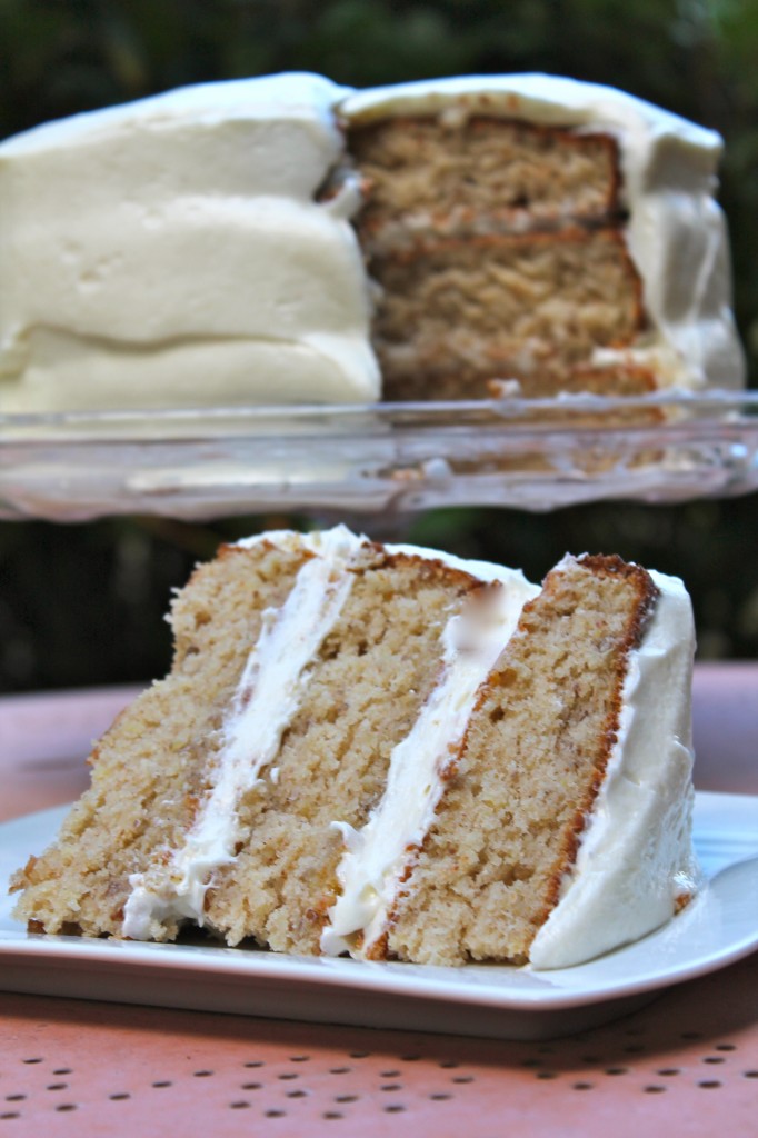 A single slice of homemade Southern hummingbird cake sits on a white plate in the foreground. In the background, a triple layer hummingbird cake with cream cheese frosting sits on a cake platter.