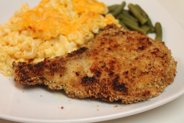 A single breaded baked pork chop is served on a plate alongside a helping of creamy mac and cheese and green beans. The breading on the chop is a crispy golden brown. Crispy baked pork chops are the perfect family meal!