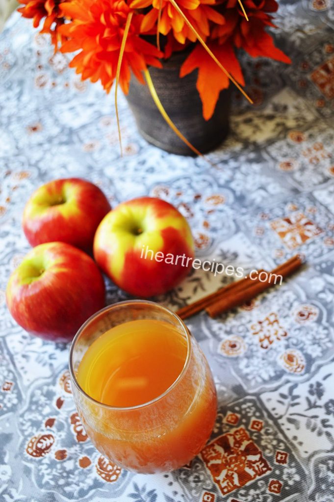 A glass filled with apple cider next to three red and yellow apples and two cinnamon sticks. A fall bouquet sits nearby.
