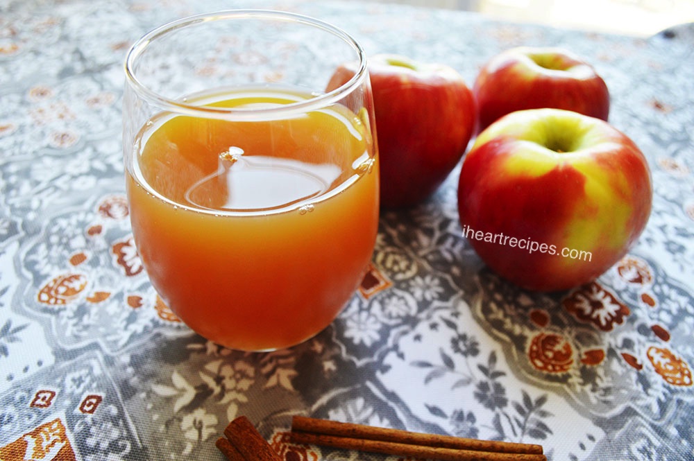 A glass filled with golden homemade apple cider. Whole apples and cinnamon sticks lay nearby.