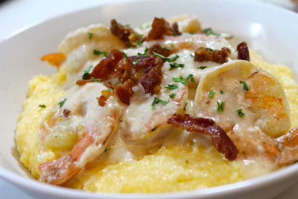 A large white porcelain bowl is filled with creamy grits topped with tender shrimp, creamy white gravy, and crispy bacon.