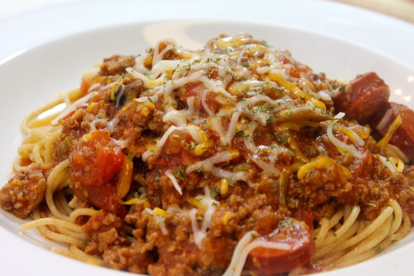 A large bowl of spaghetti and meat sauce, mixed with sliced sausage, vegetables, and shredded mozzarella cheese.