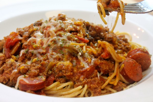 A bowl of spaghetti and sausage, made with a hearty meat sauce. The bowl of spaghetti is topped with melted shredded cheese.