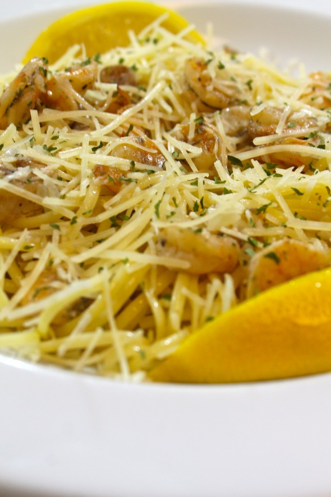 A close up of tender linguini and savory shrimp topped with grated parmesan cheese and garnished with a lemon wedge served in a white pasta dish. 