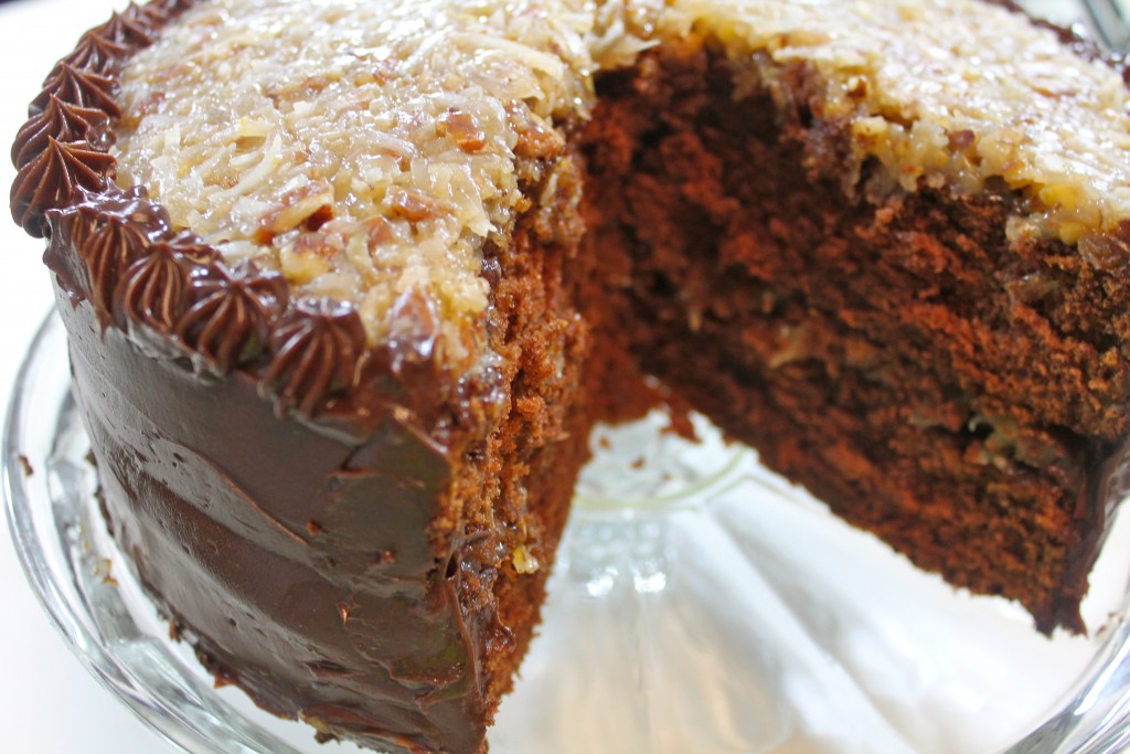 A triple layer moist chocolate cake filled with dark chocolate frosting and topped with a creamy coconut pecan frosting served on a glass cake stand. 