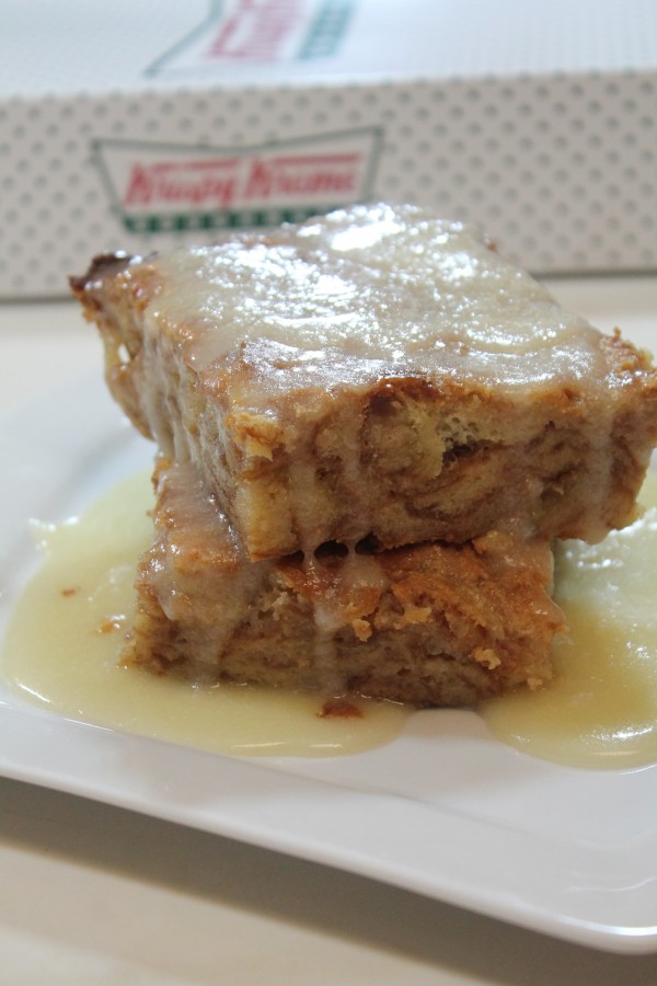 Two thick slices of Krispy Kreme bread pudding are stacked on a white plate, topped with a creamy vanilla glaze. A box of Krispy Kreme doughnuts sits in the background.