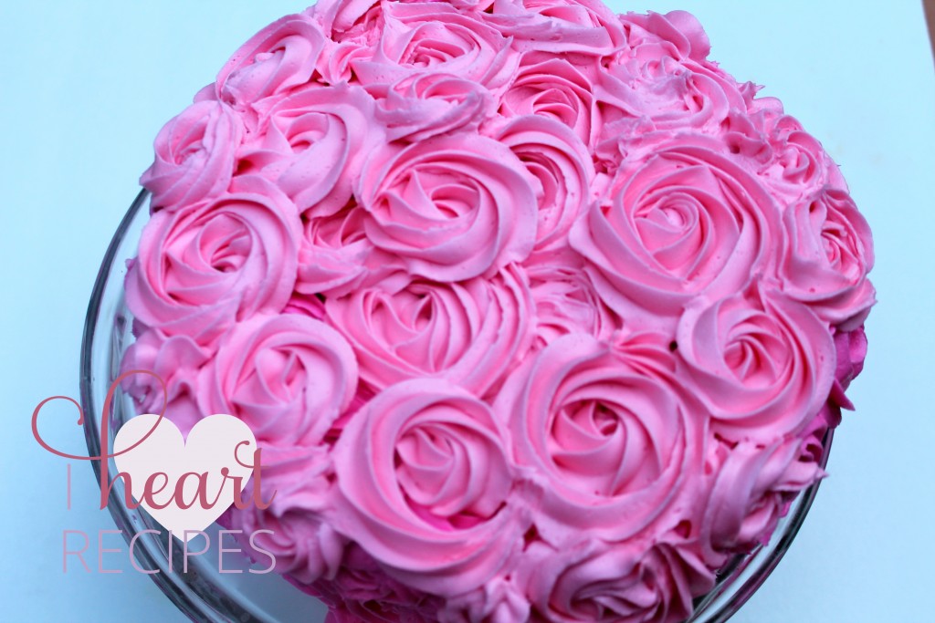 Bright pink buttercream frosting rose swirls decorate the outside of a layer cake atop a glass cake stand. 