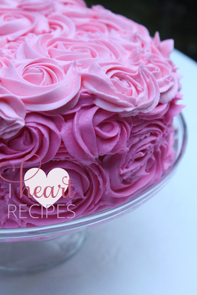 Pink ombre frosting roses decorating a  round layer cake. The cake sits atop a glass cake stand. 