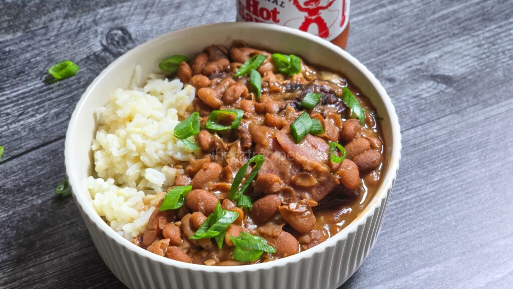 Beans and rice in a bowl with green onions on top. A bottle of hot sauce in the back of the bowl.