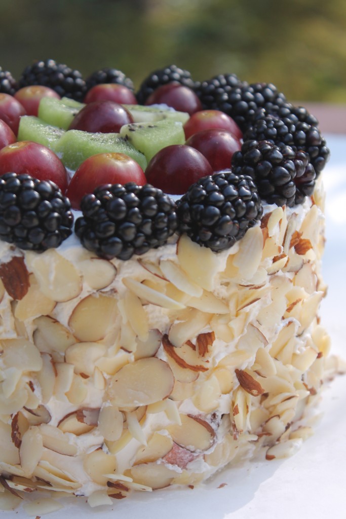 A watermelon cake covered in whipped topping and slivered almonds, with fresh blackberries, grapes, and kiwi arranged on the top of the cake.