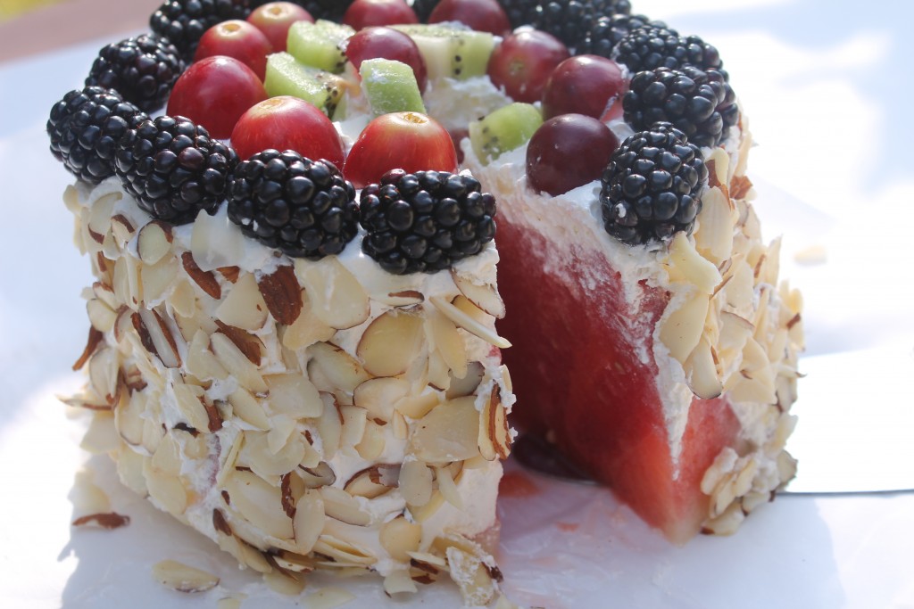 A fresh watermelon cake sits outside on a white table. Covered in sweet whipped topping, slivered almonds, and fresh fruit, this watermelon cake is the perfect summertime treat.