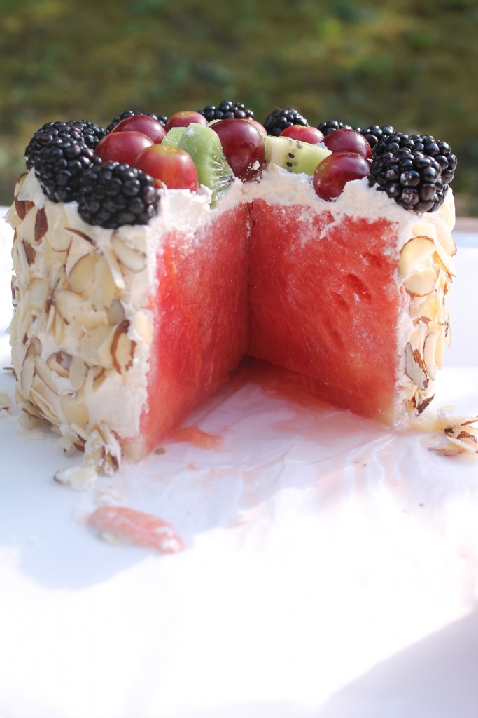 A delicious fresh watermelon cake with a large slice cut out, topped with whipped cream, blackberries, grapes, kiwi, and slivered almonds