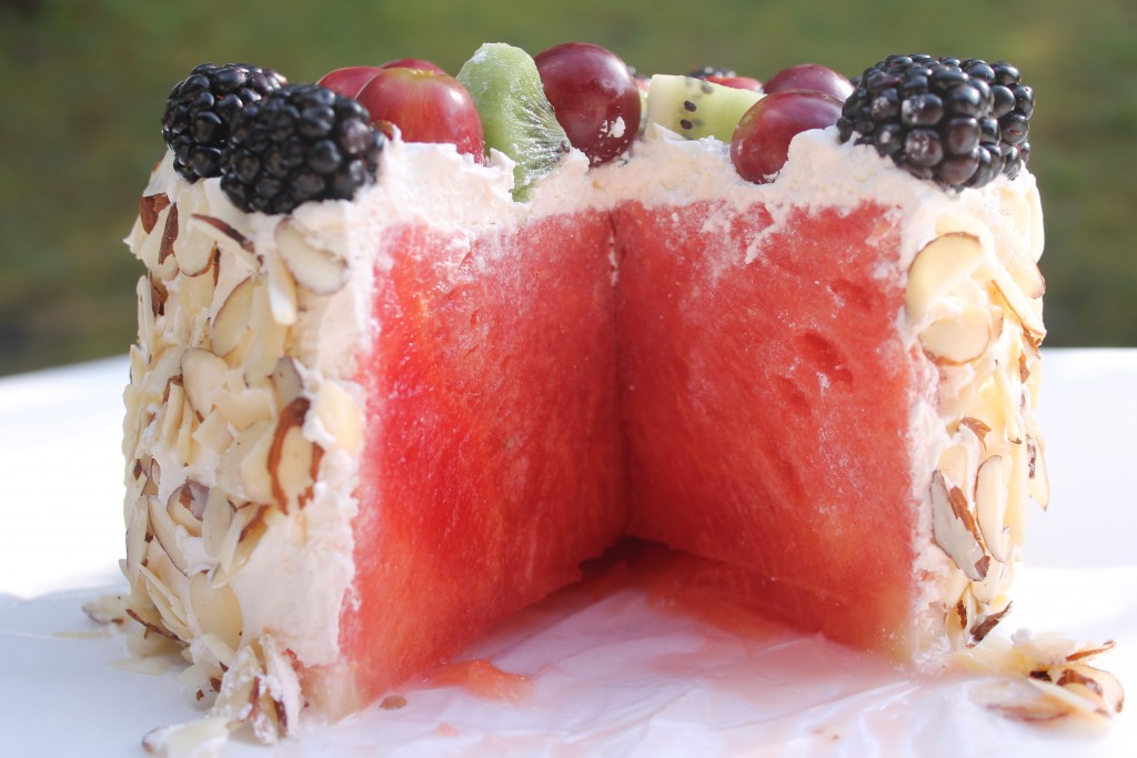 A side view of a fresh watermelon cake with a large slice cut out. The watermelon is topped with fresh whipped topping, assorted fruit, and slivered almonds.