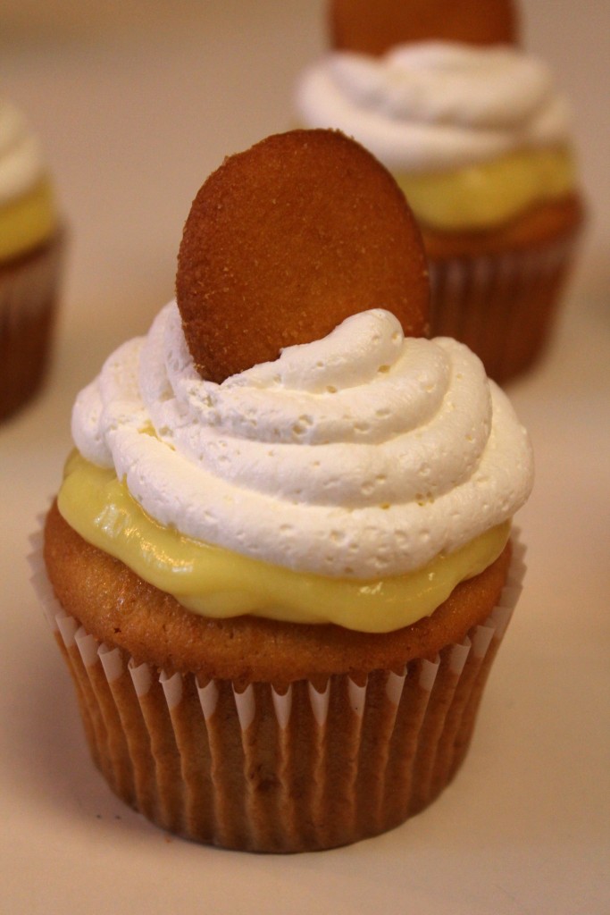 A close up of a Banana Pudding Cupcake topped with golden pudding, whipped cream and a crispy Nilla wafer.  