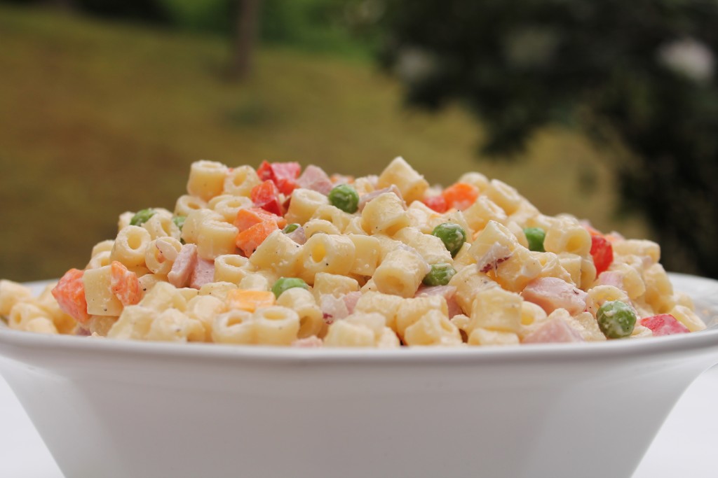 A white bowl filled with macaroni salad with colorful peas, red peppers, carrots, ham, and cheese, coated in a creamy Miracle Whip dressing.