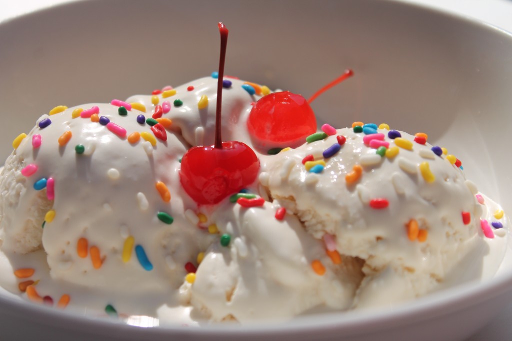 A white bowl with scoops of soft vanilla homemade ice cream topped with rainbow sprinkles and two cherries. 
