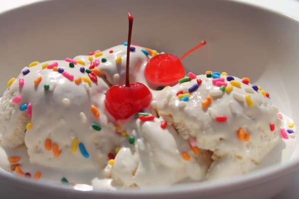 A white bowl with scoops of homemade vanilla ice cream topped with rainbow sprinkles and two cherries. 