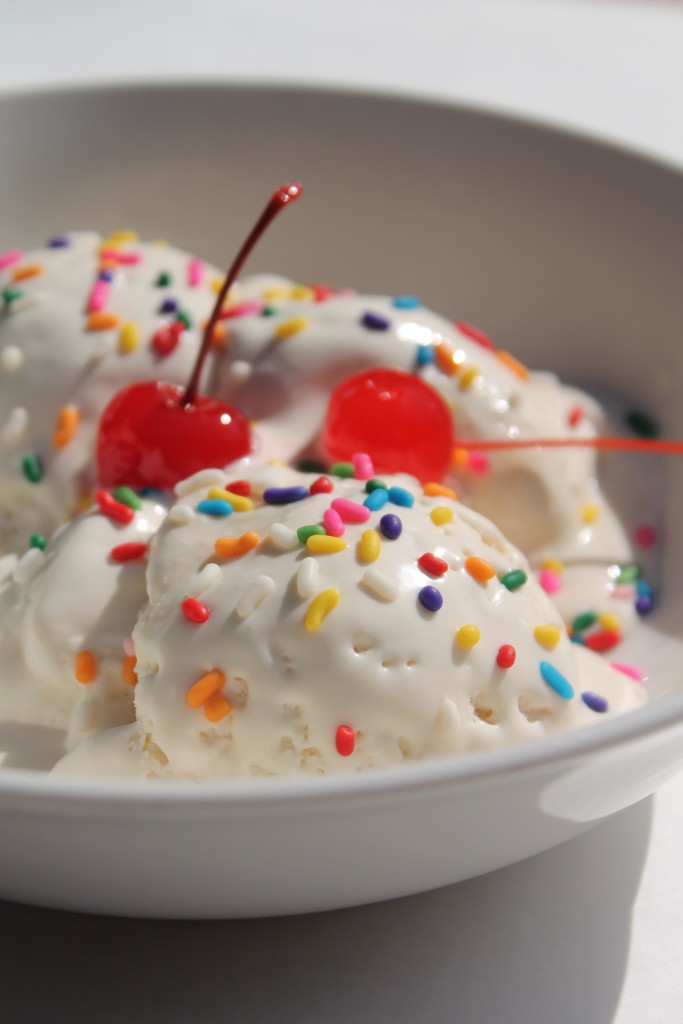 A close up of a scoop of melting vanilla ice cream topped with rainbow sprinkles and two bright red cherries. 