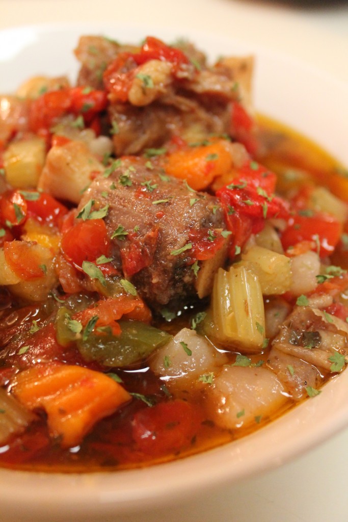 A close-up image of oxtail stew served in a white bowl. The comforting stew is made with onions, peppers, carrots, celery, potatoes, beef oxtails, and a hearty tomato broth.