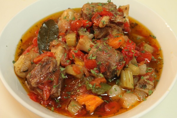 A bowlful of Slow Cooked Oxtail Stew in a red broth garnished with fresh parsley and a bayleaf. 