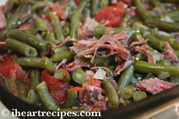 A close up image of green beans with tomatoes, smoked turkey, and onions served in a glass casserole dish.