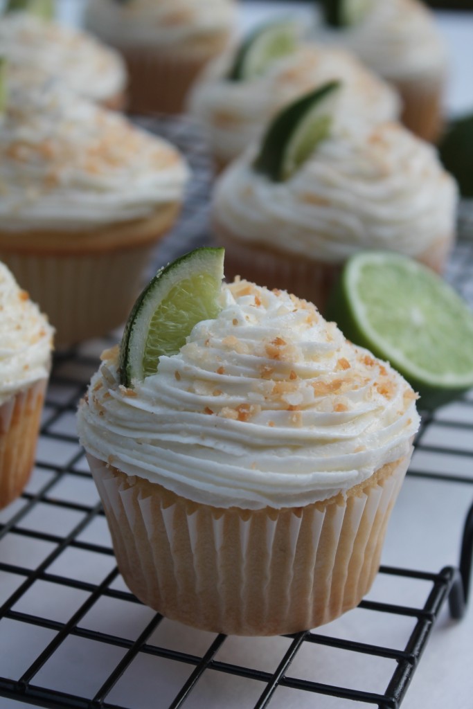 A close shot of a coconut lime cupcake garnished with a lime wedge and toasted coconut. There are cupcakes and limes in the background. 