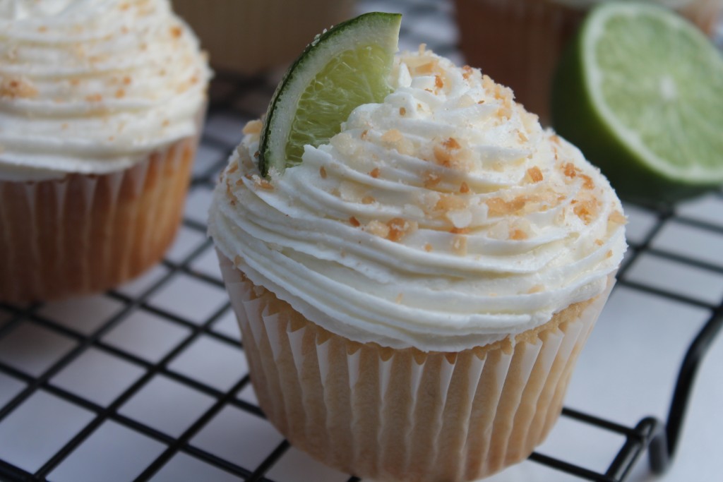 A close-up image of a coconut lime cupcake topped with a swirl of frosting, toasted coconut bits and a lime wedge. There are cupcakes in the background. 