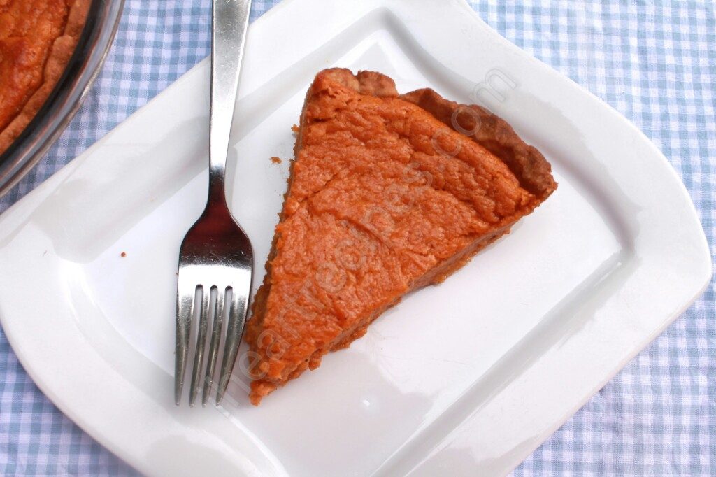 An overhead shot of a piece of sweet potato pie served on a white square plate with a silver fork. The plate sits on a light blue and white gingham patterned tablecloth.