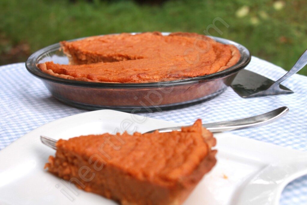 In the foreground of the image, a single slice of sweet potato pie is served on a white plate with a fork. On the gingham-covered table is the rest of the sweet potato pie in a round glass pie plate.