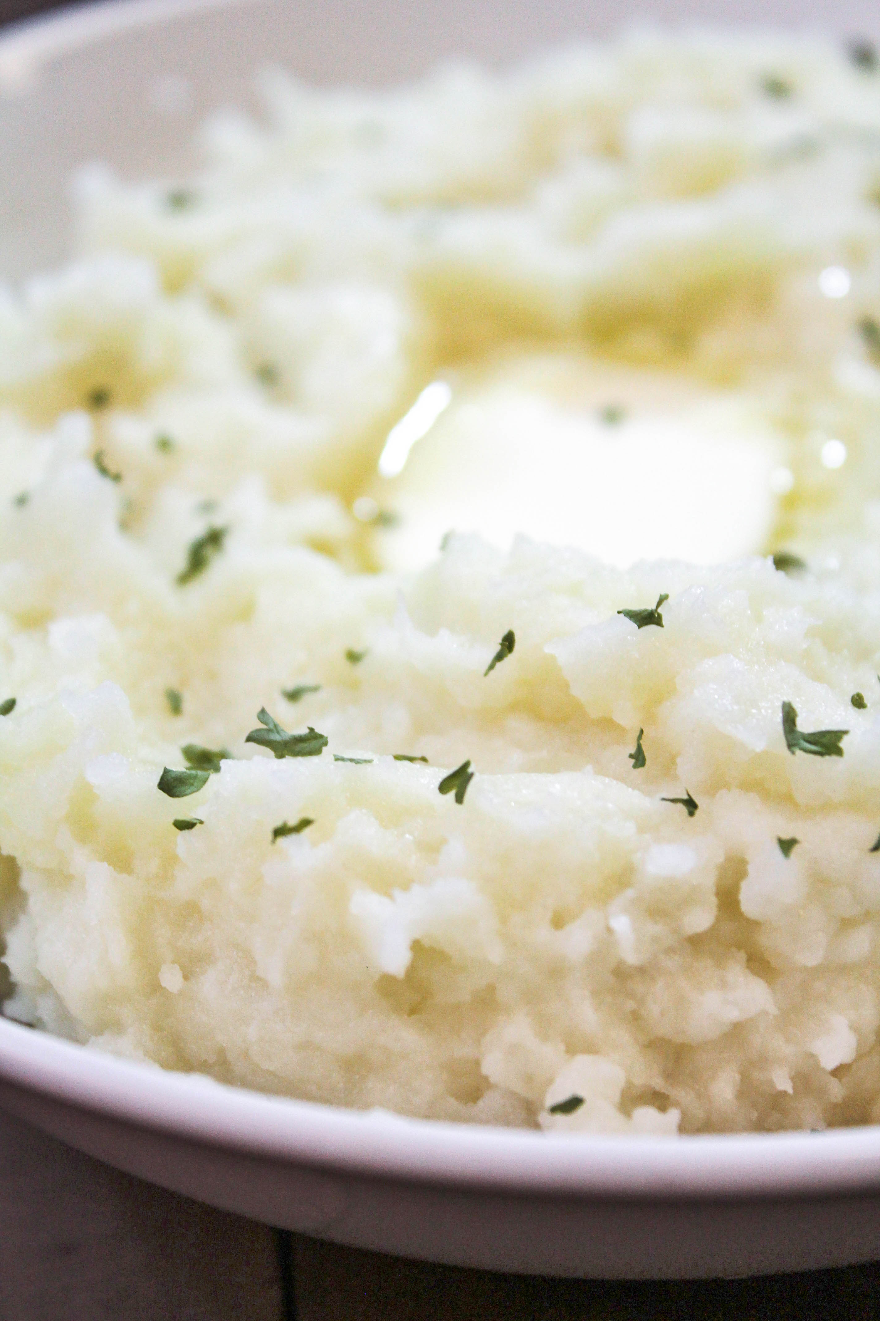 A close up image of creamy mashed potatoes, garnished with parsley flakes and a melted pat of butter.