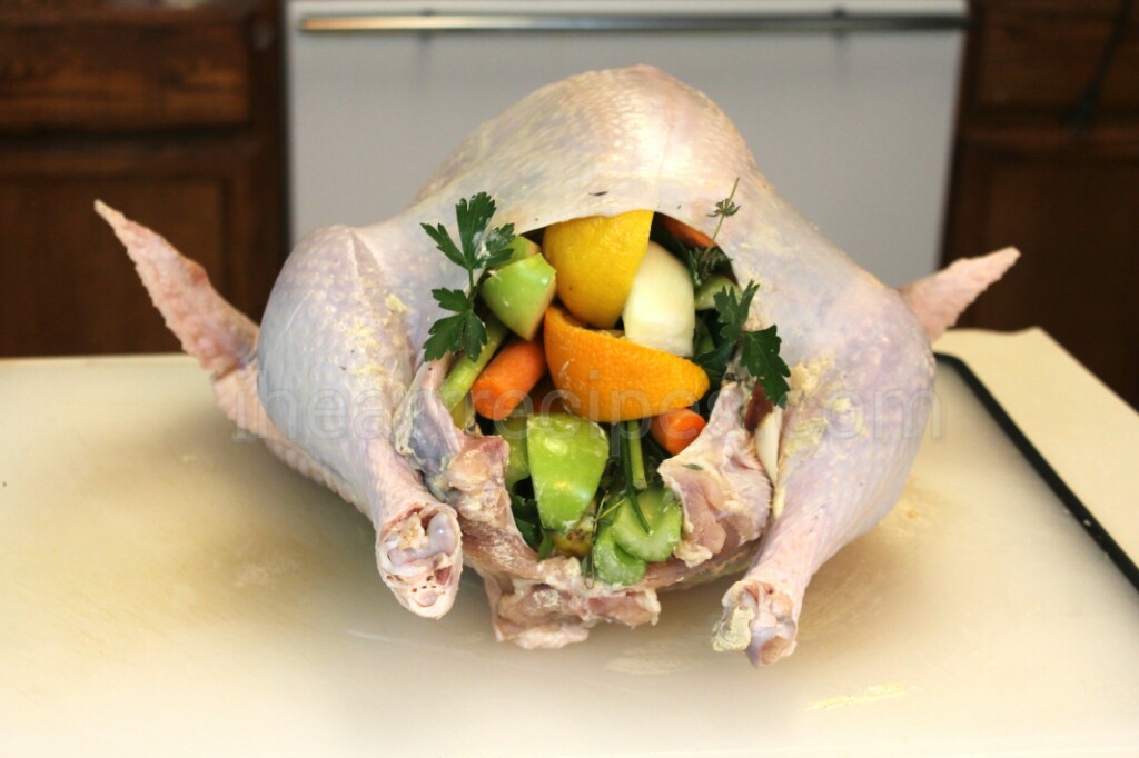 A whole raw turkey sits on a white countertop. The turkey cavity is stuffed with aromatics like apples, oranges, carrots, and fresh herbs.