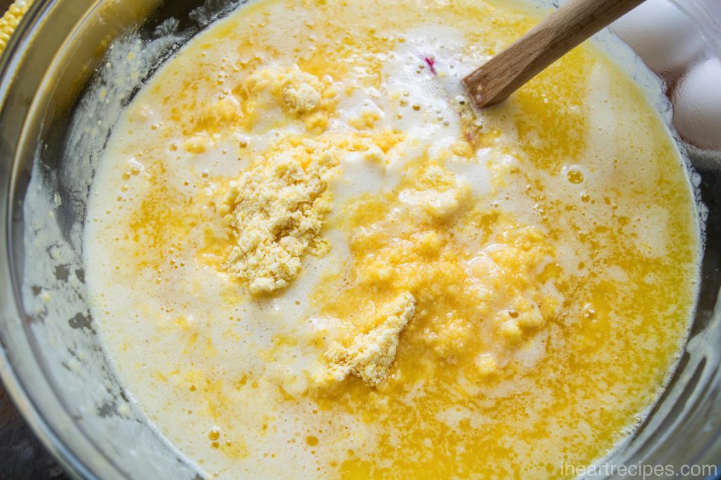The ingredients for corn pudding casserole—creamed corn, cornbread mix, sour cream, sugar, and butter—mixed with a wooden spoon in a large glass mixing bowl. 