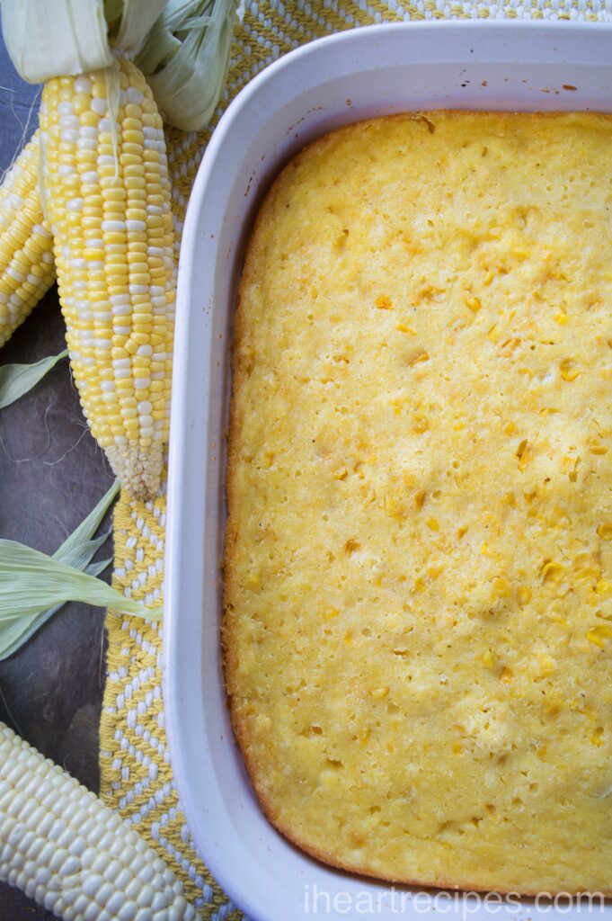 A white ceramic casserole dish filled with fresh-baked corn casserole. Ears of shucked white and yellow corn cobs sit alongside the casserole dish.