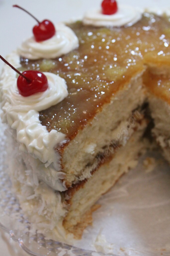 A close-up look at a layered coconut pineapple cake topped with a sweet pineapple filling, frosting, and cherry garnish. A large slice is cut from the whole cake, revealing the layered inside.