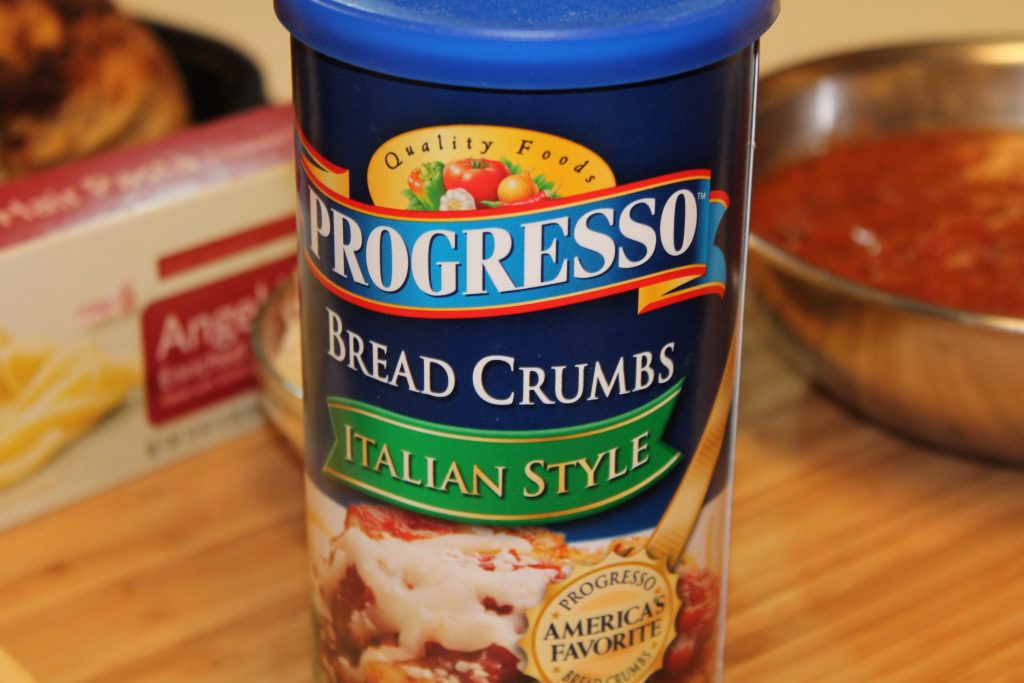 A close-up of a blue container of Progresso Italian Style bread crumbs. A whole chicken, a box of pasta and a bowl of red sauce are in the background. 