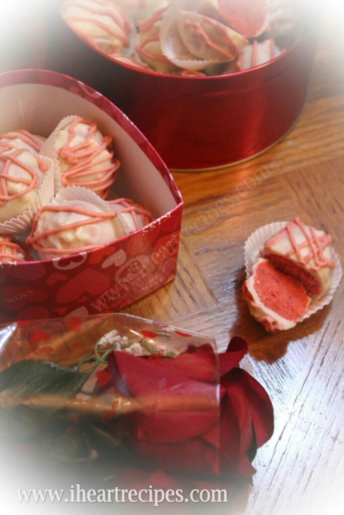 A halved pink Strawberries and Cream Cake Truffle on a wooden table next to two decorative containers filled with truffles. A red rose lays nearby.